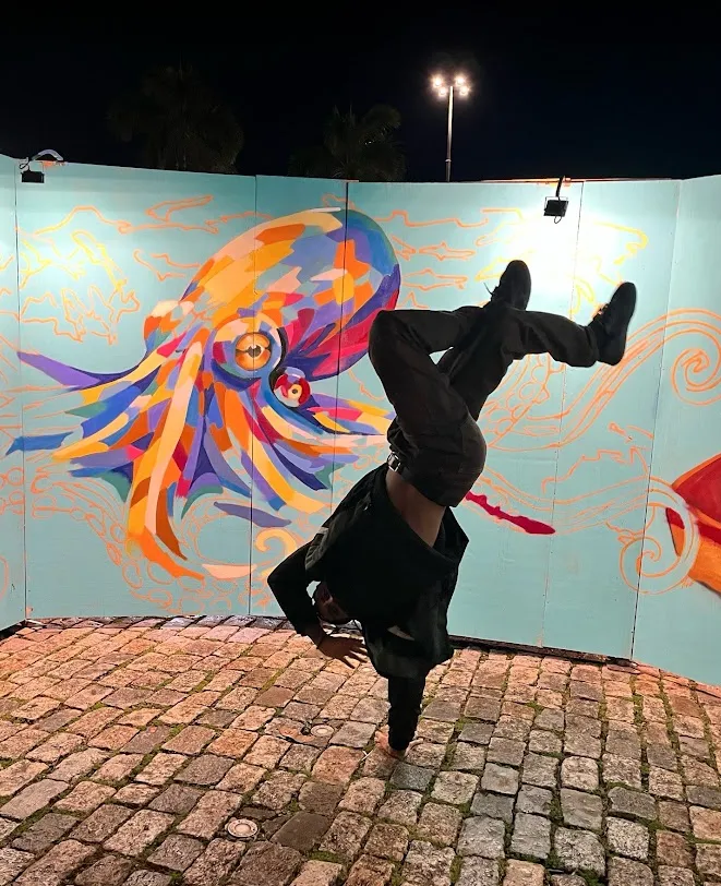 Renan doing a one-hand freeze upside down at night, in front of a mural of a colorful octopus